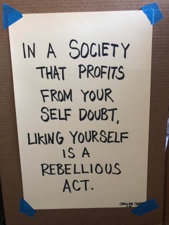 A poster by Caroline Caldwell, which reads, in a society that profits from your self doubt, liking yourself is a rebellious act.